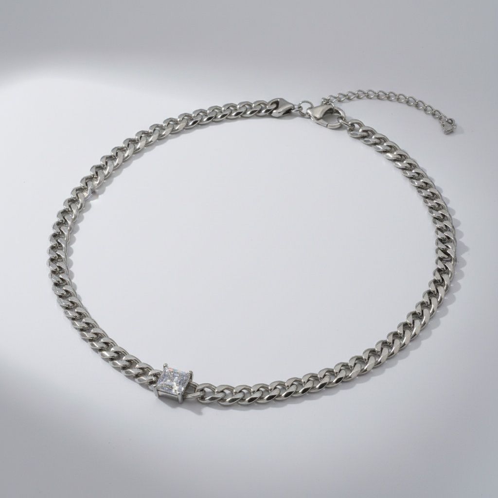 Overhead flat lay of an 8mm stainless steel Cuban curb chain necklace in silver arranged in a full circle on a light grey surface. A single princess cut square cubic zirconia stone is bezel-set at the bottom center of the chain. A heavy duty lobster clasp and 2-inch extender chain are visible at the top right.