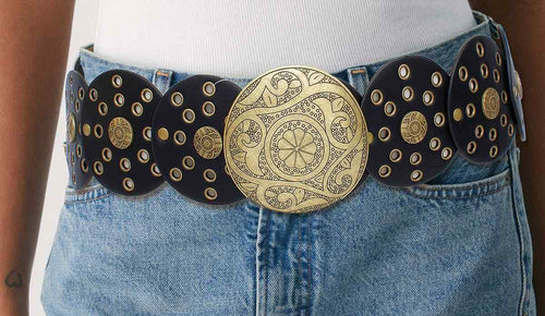 Black vegan faux leather wide western concho belt worn on woman's waist over white ribbed top and light blue denim mini skirt, large engraved distressed gold round concho buckle with floral paisley pattern, black disc panels with gold grommet punch-out details