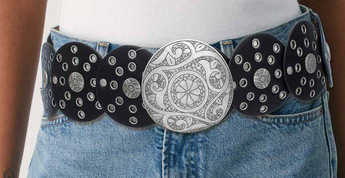 Black vegan faux leather wide western concho belt worn on woman's waist over white ribbed top and light blue denim mini skirt, large engraved distressed silver round concho buckle with floral paisley pattern, black disc panels with silver grommet punch-out details