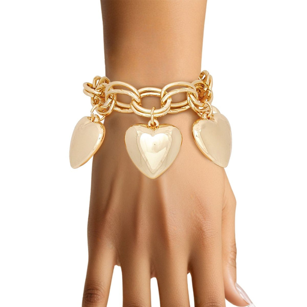 Close-up of a woman's wrist and hand with light skin and natural nails against a white background, wearing a chunky gold tone double oval link chain bracelet with five oversized puffy hollow heart charms dangling from the links. The heart charms have a smooth polished gold finish and appear cream-toned against the skin in the light.