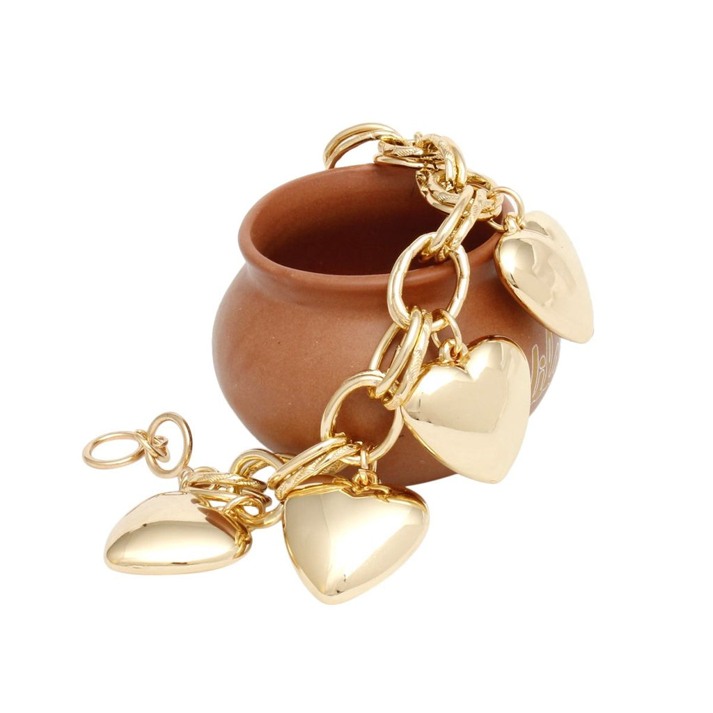 Angled lifestyle shot of a chunky gold tone double oval link chain charm bracelet with five oversized puffy hollow polished gold heart charms draped over and around a small round terracotta clay pot on a white background. The toggle bar closure and ring are visible at the left side.