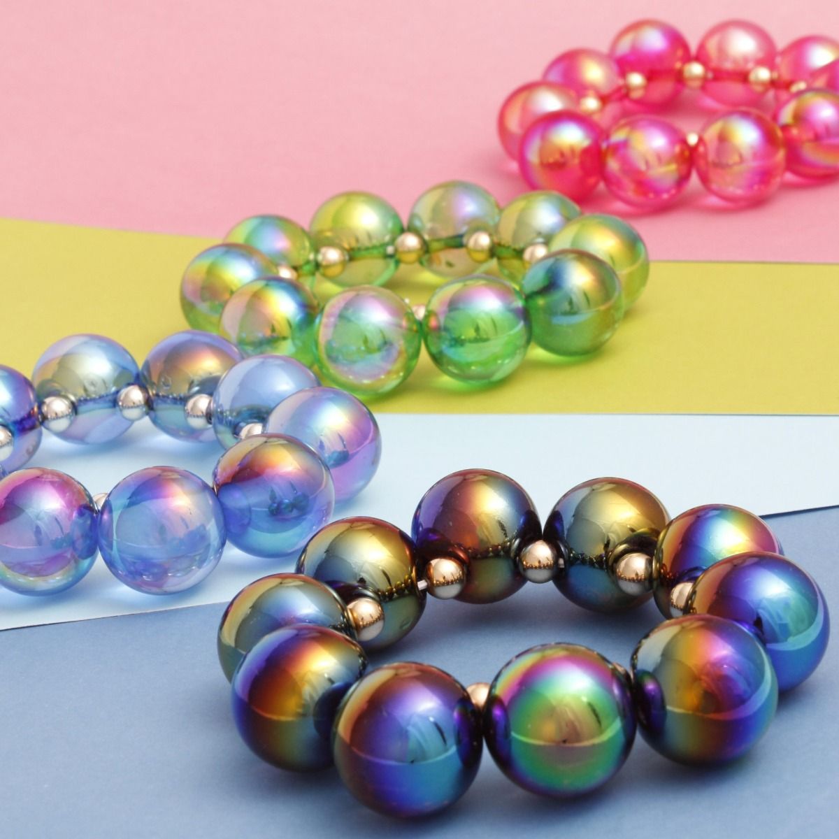 Black rainbow iridescent bubble bead bracelet in the foreground with blue, green, and pink bead bracelets behind it on pastel-colored surfaces