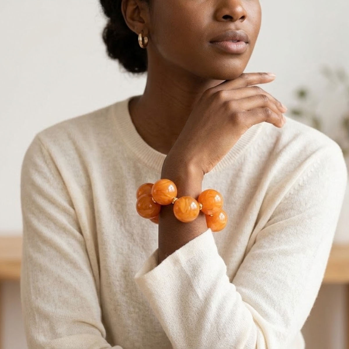 Model wearing an orange swirled bubble bead bracelet with a cream sweater, her hand raised near her chin in a softly blurred indoor setting