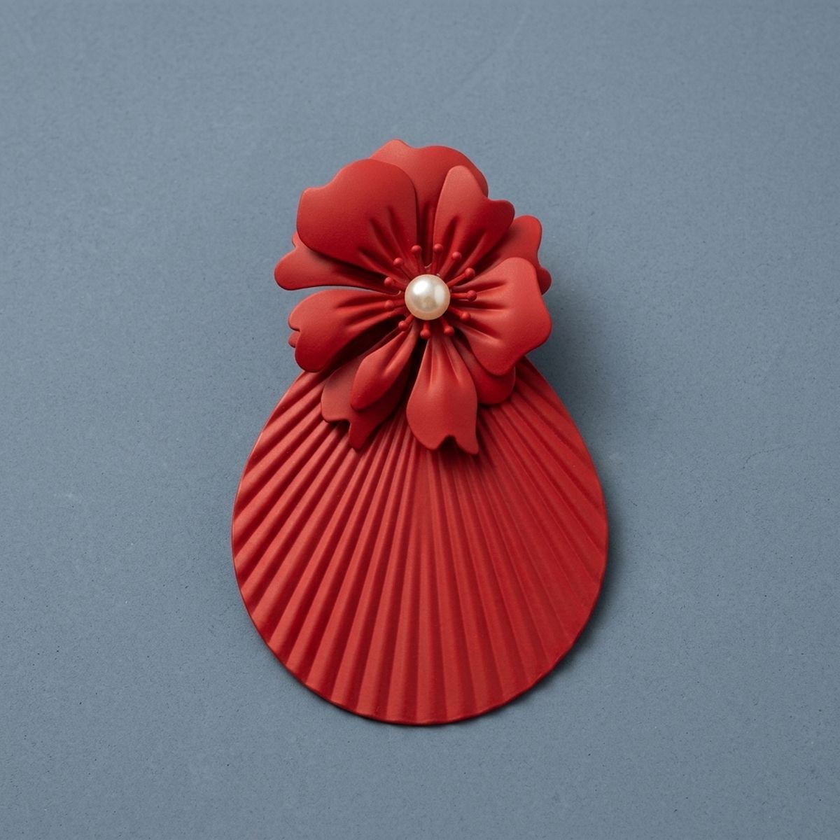 Single oversized matte red statement earring with layered flower petals, pearl-look center, and pleated fan-shaped teardrop drop on a slate blue background