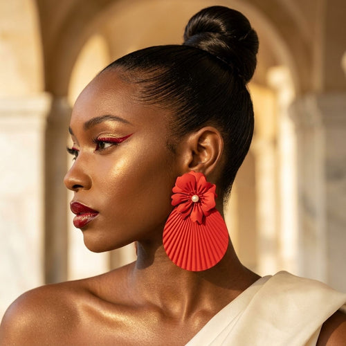Black woman in side profile wearing an oversized matte red floral statement earring with pearl-look center and pleated teardrop drop, styled with a high bun and white one-shoulder top against a warm arched background