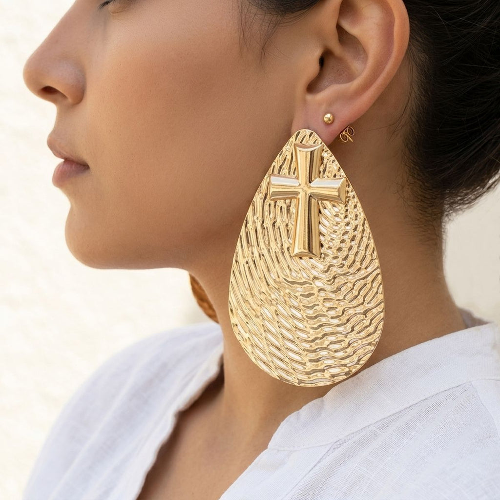 Model wearing an oversized gold-tone textured teardrop stud earring with raised cross detail