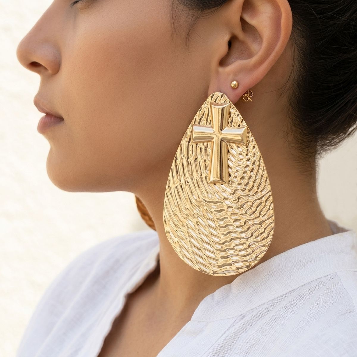 Model wearing an oversized gold-tone textured teardrop stud earring with raised cross detail