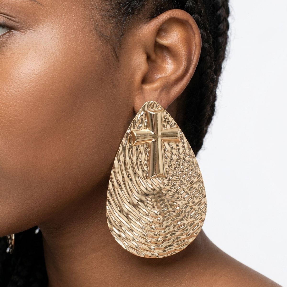 Model wearing an oversized gold-tone teardrop earring with ripple texture and raised cross emblem