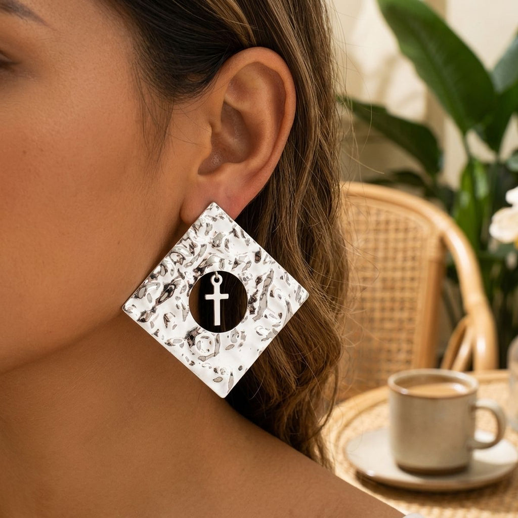 Model wearing an oversized hammered silver-tone diamond-shaped stud earring with a circular cutout and dangling cross charm in a café setting