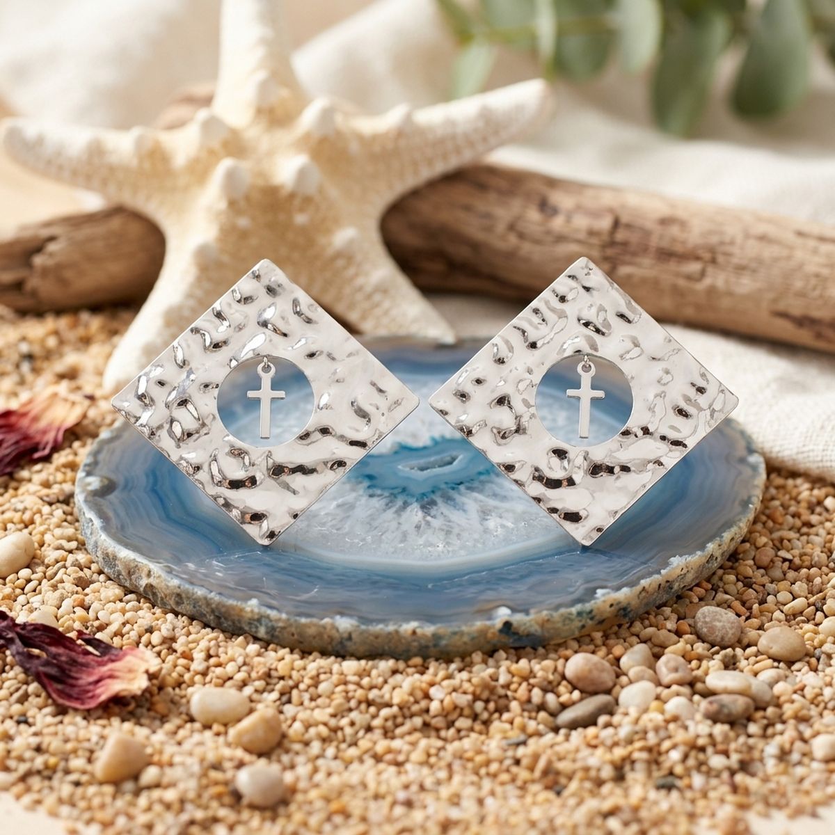 Pair of hammered silver-tone diamond-shaped stud earrings with circular cutouts and cross charms displayed on a blue stone with beach décor