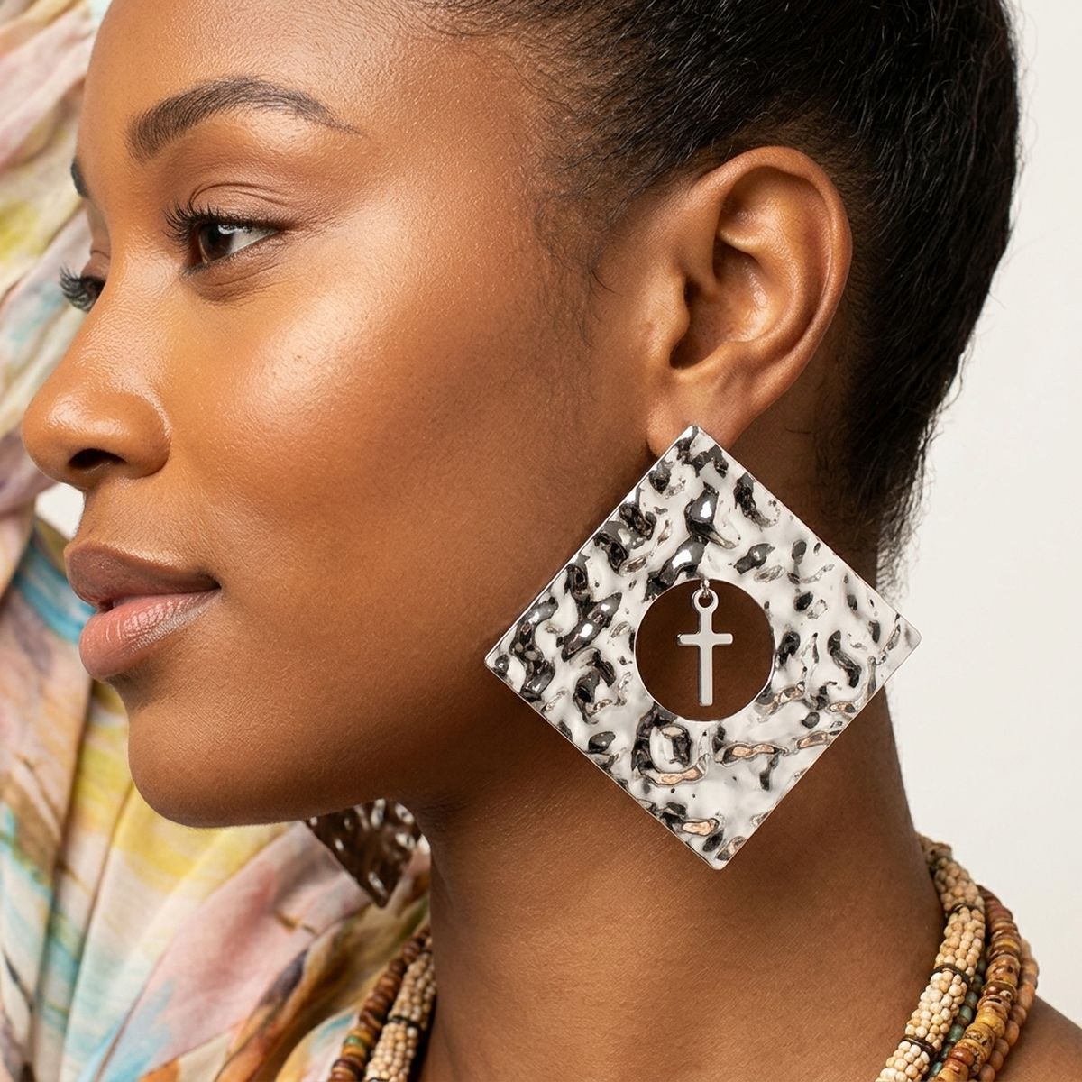 Model wearing an oversized hammered silver-tone diamond stud earring with a round center cutout and dangling cross charm against a colorful background