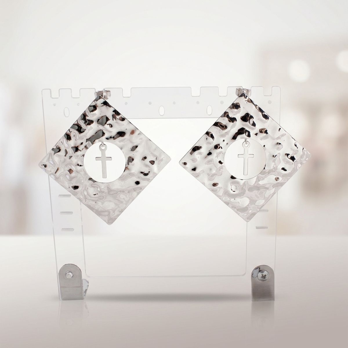 Pair of oversized hammered silver-tone diamond-shaped stud earrings with round center cutouts and dangling cross charms on a clear display stand