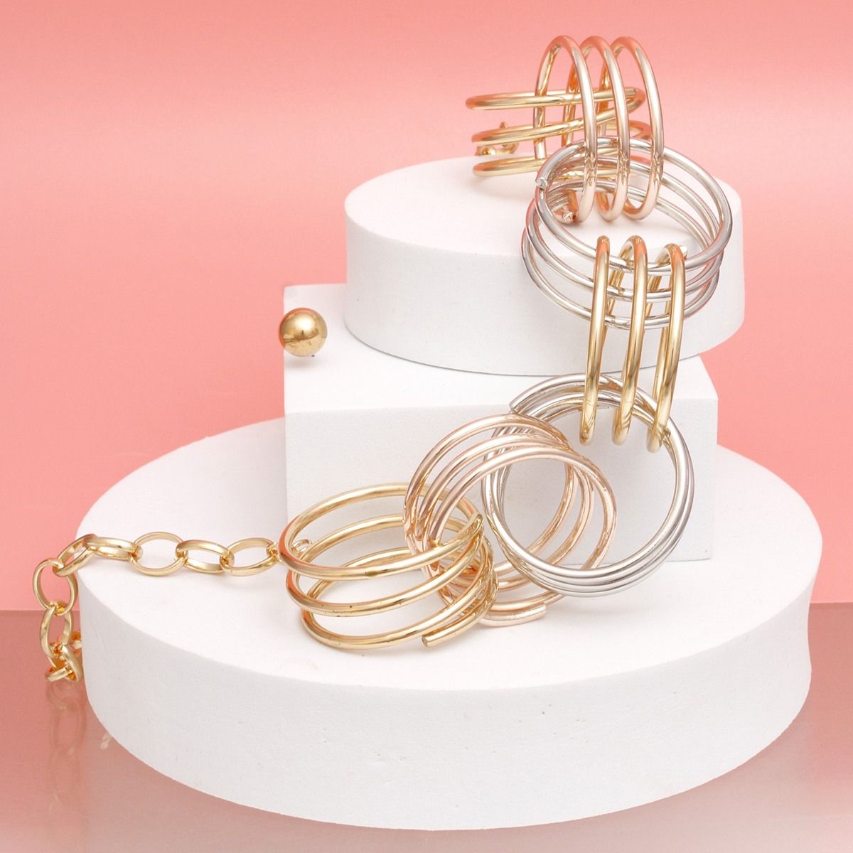Angled wide shot of a mixed metal multi-wire open ring collar necklace draped across two white cylindrical tiered display stands against a coral pink background. The oversized multi-wire open rings in gold, rose gold, and silver tone are visible, along with the gold tone oval link chain and extender. A small gold tone ball stud earring is visible on the lower left stand.