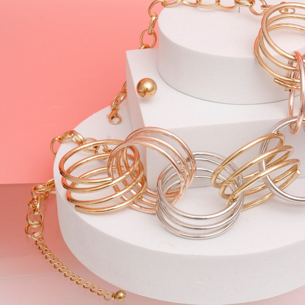 Close-up angled shot of the collar section of a mixed metal multi-wire open ring collar necklace draped across white cylindrical tiered display stands against a coral pink background. The interlocking oversized multi-wire open rings in gold, rose gold, and silver tone are shown in detail, with the gold tone oval link chain and 3-inch extender chain visible at the lower left.
