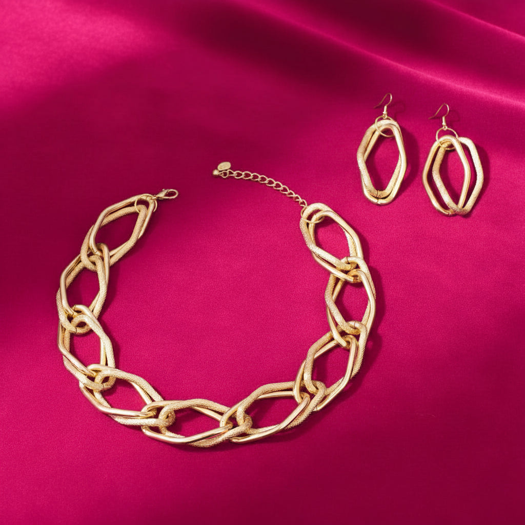 Overhead flat lay of an oversized diamond cut geometric open link chain necklace in matte gold tone arranged in a circle on a magenta pink satin fabric background, with a lobster clasp and extender chain visible at the top left. Two matching open geometric diamond-shaped drop earrings with fish hook backs are displayed to the upper right.