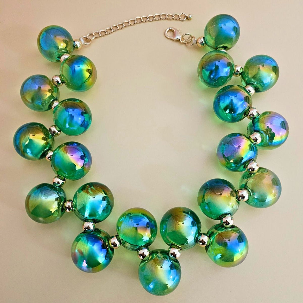 Close-up of green iridescent bubble bead necklace with blue, purple, and gold reflections, metal spacers, lobster clasp, and extender on a beige background
