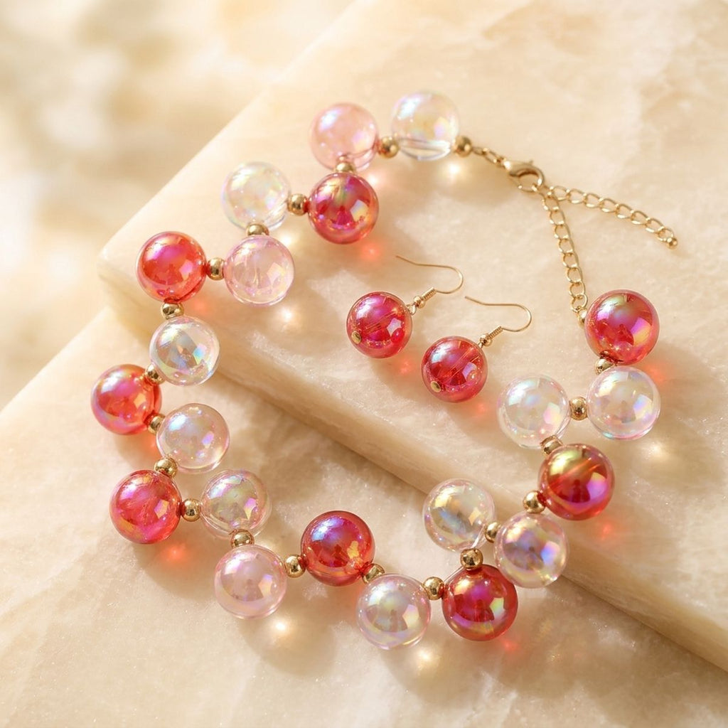 Red and pink iridescent bubble-bead necklace with gold spacers and matching drop earrings arranged on cream fabric