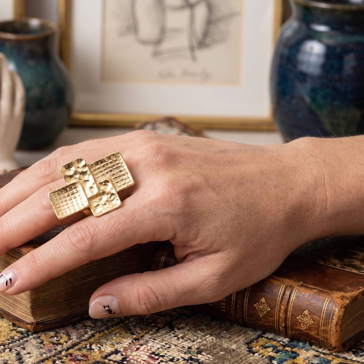 Woman’s hand wearing a chunky gold-tone stretch ring with four overlapping hammered and grid-textured geometric panels over vintage books