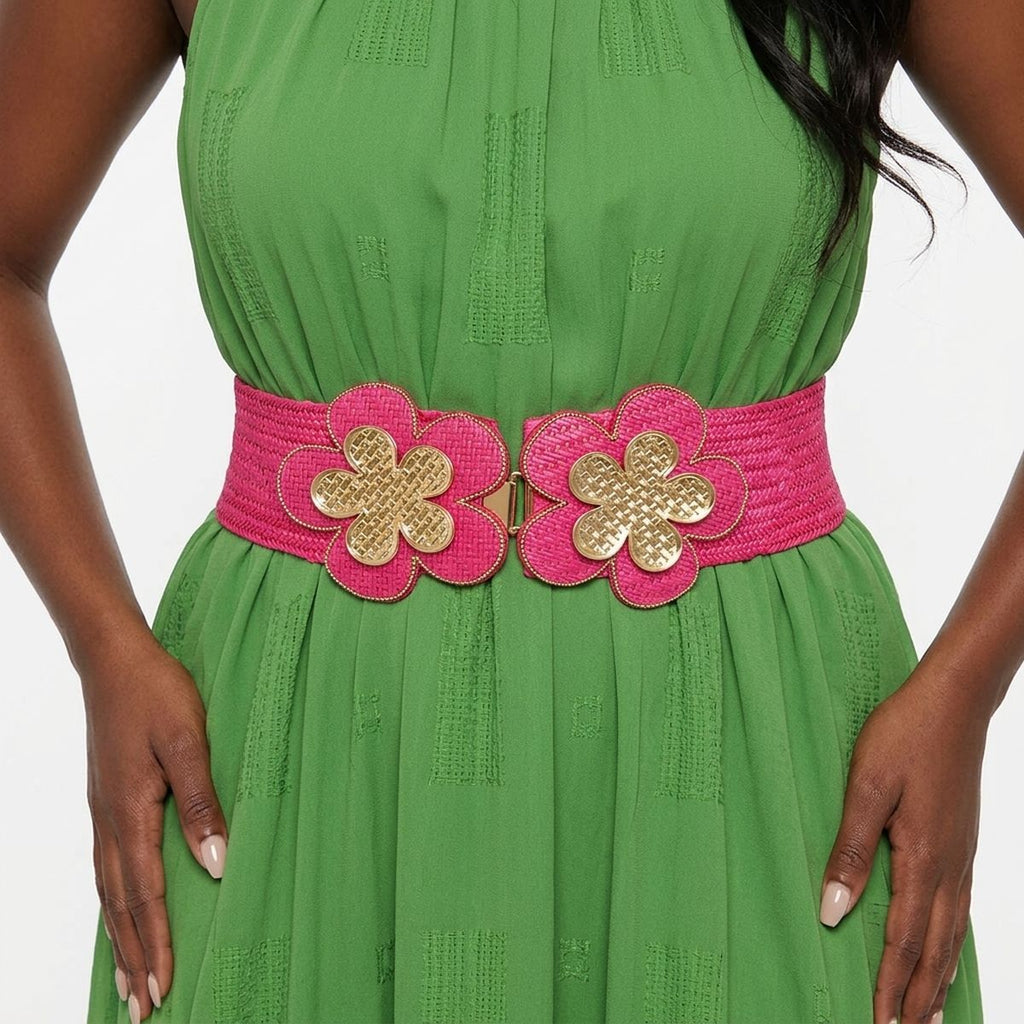 Closeup of fuchsia wide woven raffia stretch belt worn on plus size woman's waist over green halter neck jacquard maxi dress, double gold basket-weave flower hook closure at center showing two five-petal flowers with gold crosshatch texture centers and fuchsia raffia petal surrounds with gold beaded trim edging, white background
