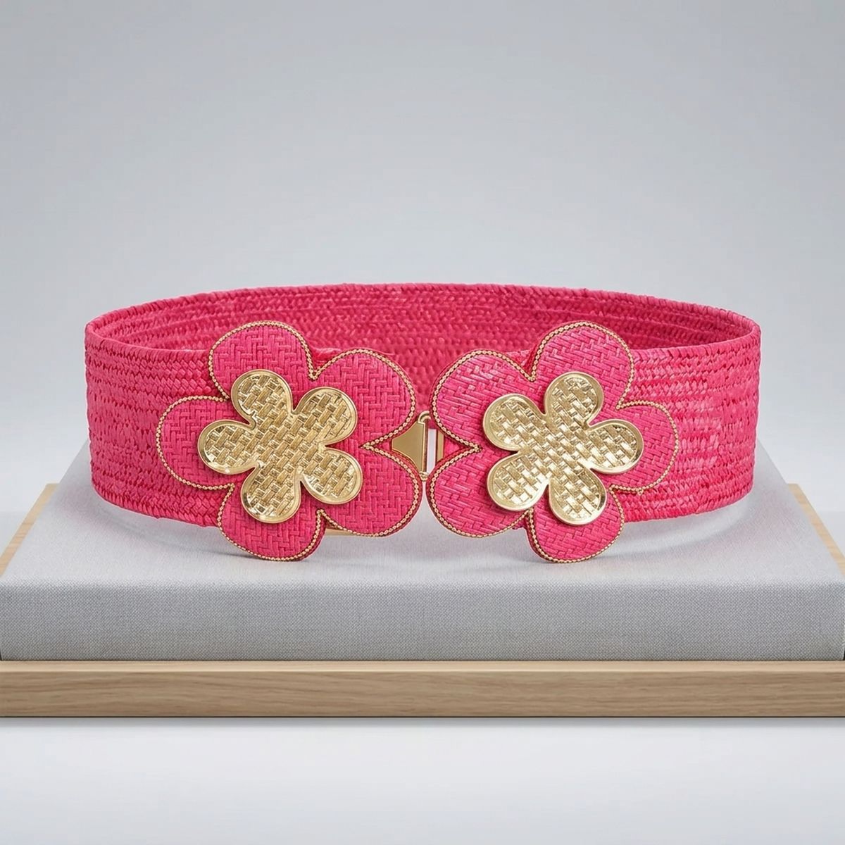 Fuchsia wide woven raffia stretch belt displayed in oval loop on gray linen surface, double gold basket-weave flower hook closure at center showing two five-petal flowers with gold crosshatch texture centers, fuchsia raffia petal surrounds with gold beaded trim edging and center hook mechanism