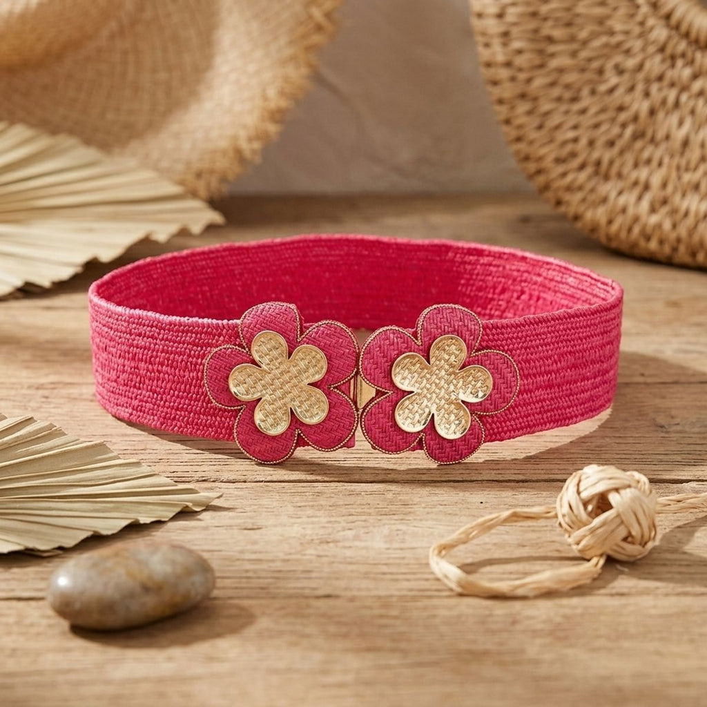 Fuchsia wide woven raffia stretch belt displayed in oval loop on rustic wooden surface, double gold basket-weave flower hook closure at center showing two five-petal flowers with gold crosshatch texture centers and fuchsia raffia petal surrounds with gold beaded trim edging, surrounded by dried palm leaves, smooth river stone, coiled natural raffia twine, and woven straw hat and basket in background