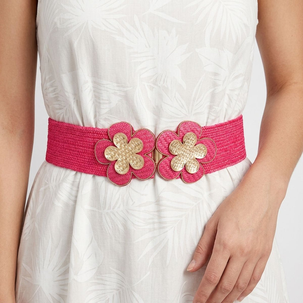 Closeup of fuchsia wide woven raffia stretch belt worn on woman's waist over white tropical leaf print sleeveless fit-and-flare dress, double gold basket-weave flower hook closure at center showing two five-petal flowers with gold crosshatch texture centers and fuchsia raffia petal surrounds with gold beaded trim edging, white background