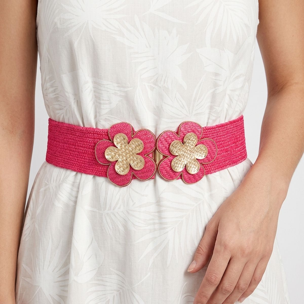 Closeup of fuchsia wide woven raffia stretch belt worn on woman's waist over white tropical leaf print sleeveless fit-and-flare dress, double gold basket-weave flower hook closure at center showing two five-petal flowers with gold crosshatch texture centers and fuchsia raffia petal surrounds with gold beaded trim edging, white background