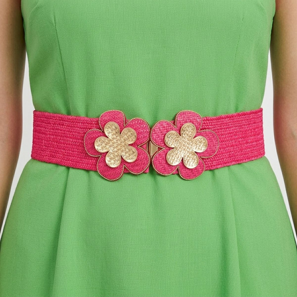 Closeup of fuchsia wide woven raffia stretch belt worn on woman's waist over green sleeveless A-line dress, double gold basket-weave flower hook closure at center showing two five-petal flowers with gold crosshatch texture centers and fuchsia raffia petal surrounds with gold beaded trim edging, white background
