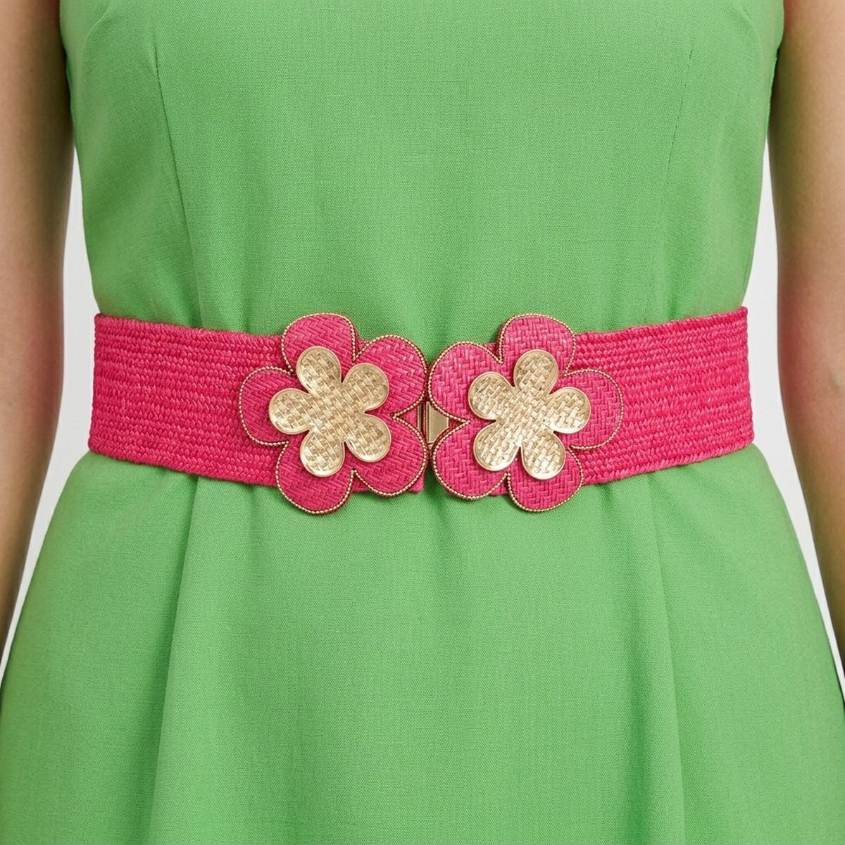 Closeup of fuchsia wide woven raffia stretch belt worn on woman's waist over green sleeveless A-line dress, double gold basket-weave flower hook closure at center showing two five-petal flowers with gold crosshatch texture centers and fuchsia raffia petal surrounds with gold beaded trim edging, white background
