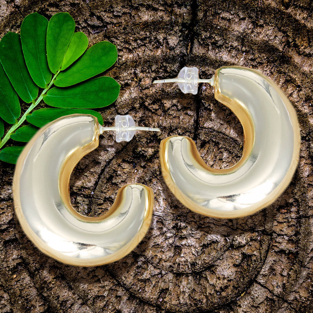 Pair of 14K gold plated chunky puffy hoop earrings with clear push back closures displayed on rustic wood surface with green tropical leaf, showing thick gold tube hoop body and reflective finish