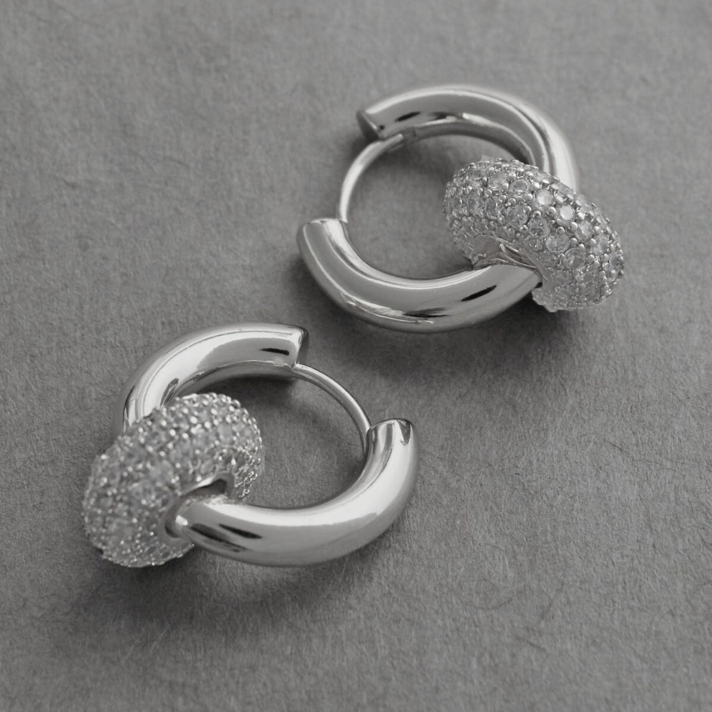 Two 14K white gold plated CZ pavé ring huggie hoop earrings displayed on gray textured surface, close-up showing polished silver hoop body and fully pavé-set cubic zirconia ring charm detail
