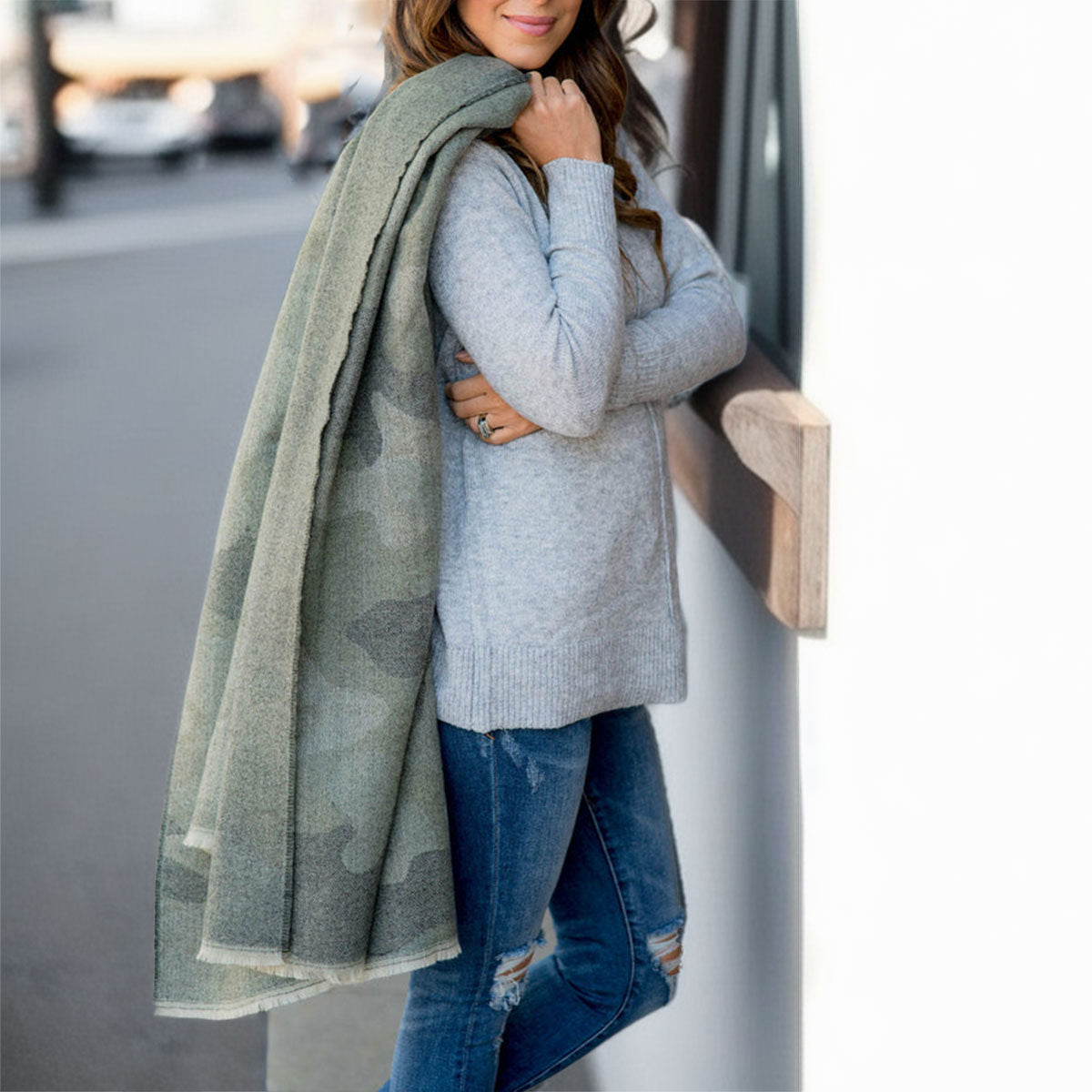 Model wearing a gray sweater and distressed blue jeans with a long faded gray camouflage scarf draped over one shoulder beside a white wall