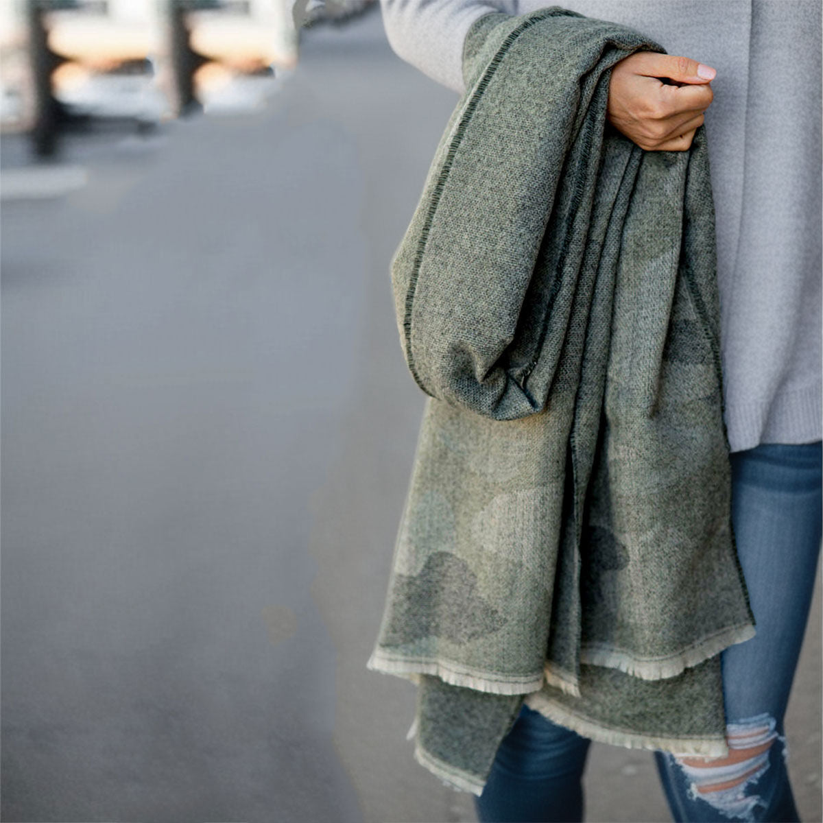 Close-up of a faded gray and charcoal camouflage knit scarf with pale fringe, held over a model’s arm beside a gray sweater and distressed blue jeans