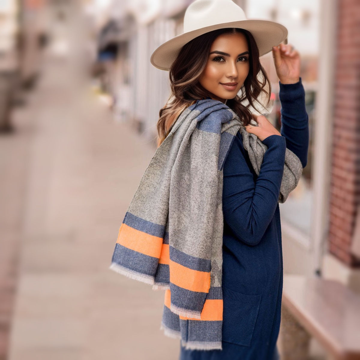 Model in a navy outfit and cream wide-brim hat carrying a gray camouflage scarf over her shoulder, with blue and orange stripes visible against a blurred city background
