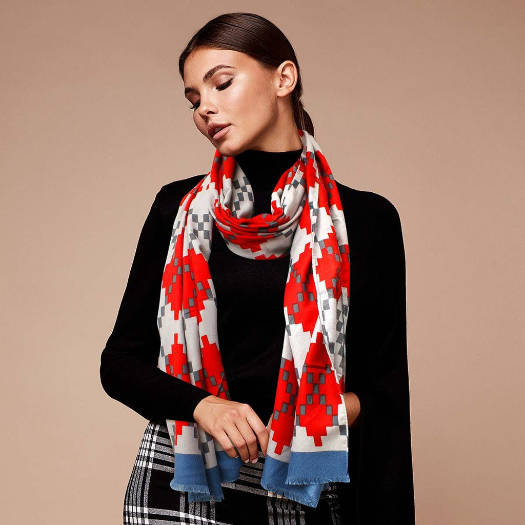 Model wearing a red pixel argyle scarf looped over a black high-neck top, with blue borders hanging above black-and-white plaid trousers against a tan background