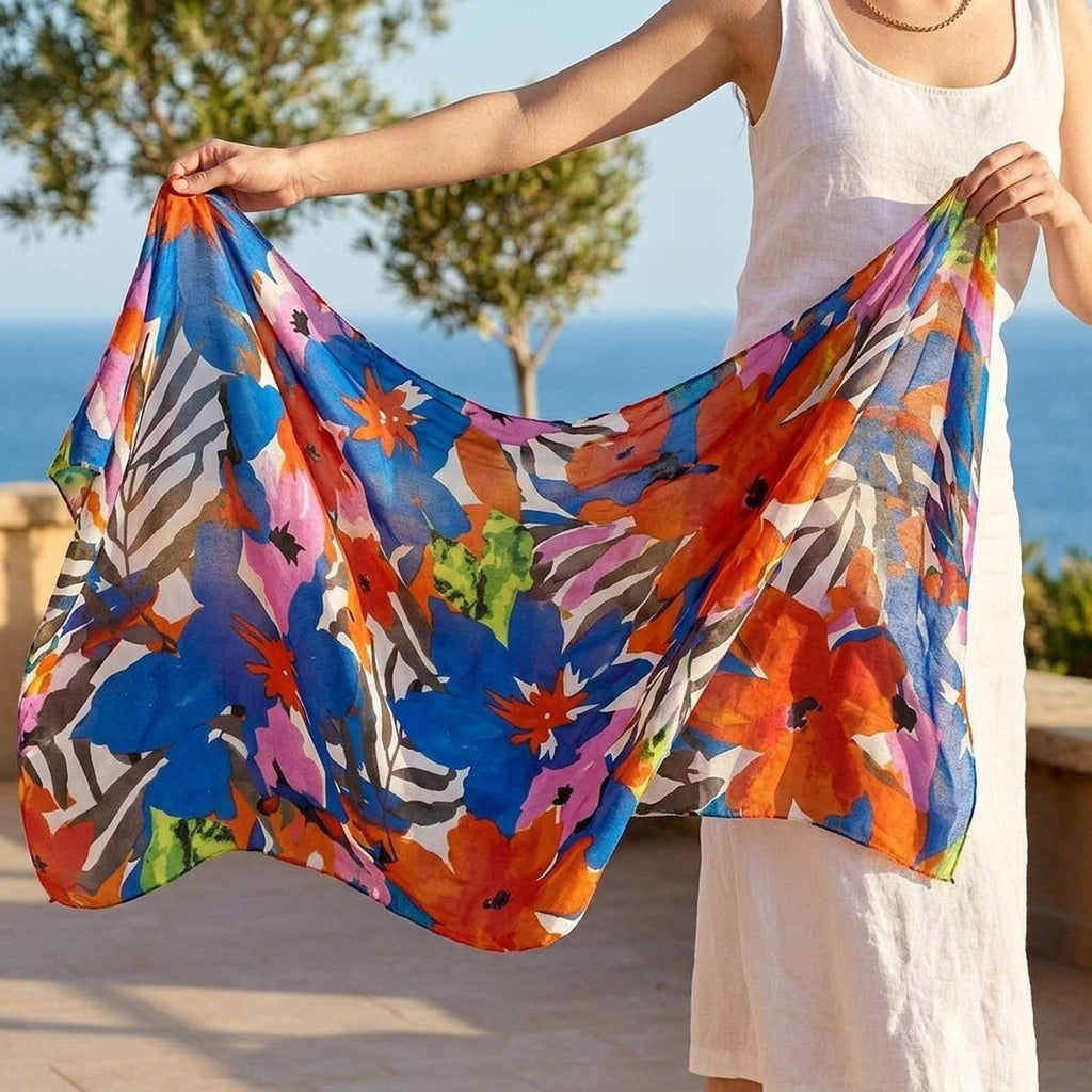 Model in a white sleeveless dress holding a blue-and-orange watercolor floral wrap open on a sunny terrace overlooking the sea