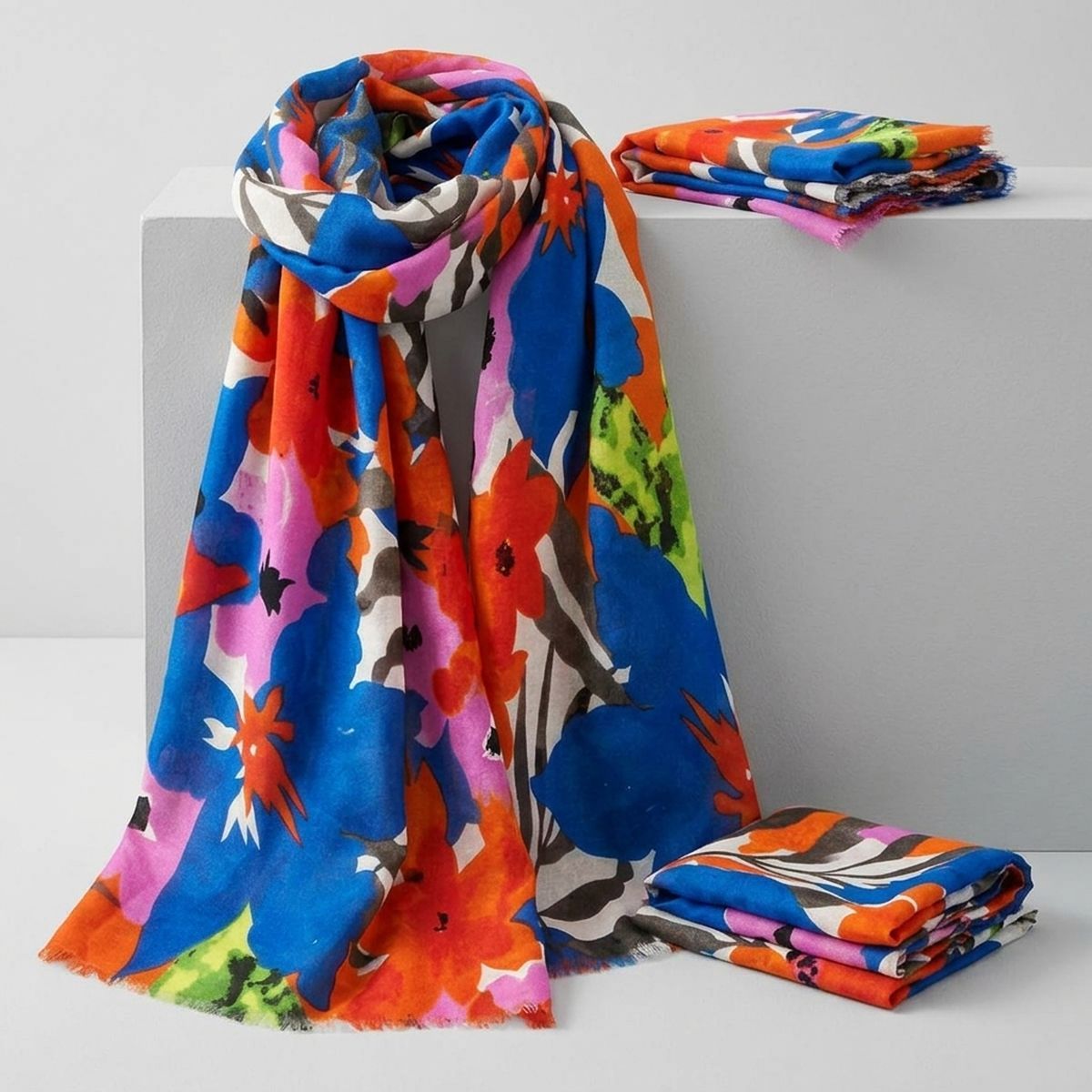 Blue-and-orange watercolor floral scarf draped over a pale display block beside folded matching scarves, with its fine frayed ends visible