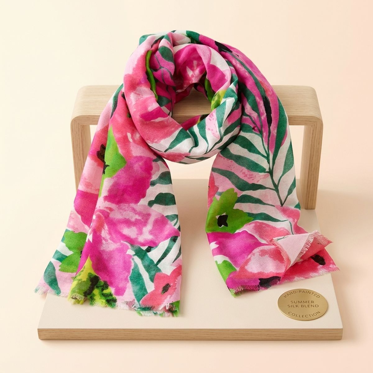 Pink-and-green watercolor floral scarf looped over a small wooden stand, showing dark flower centers and fine frayed ends on a cream display board