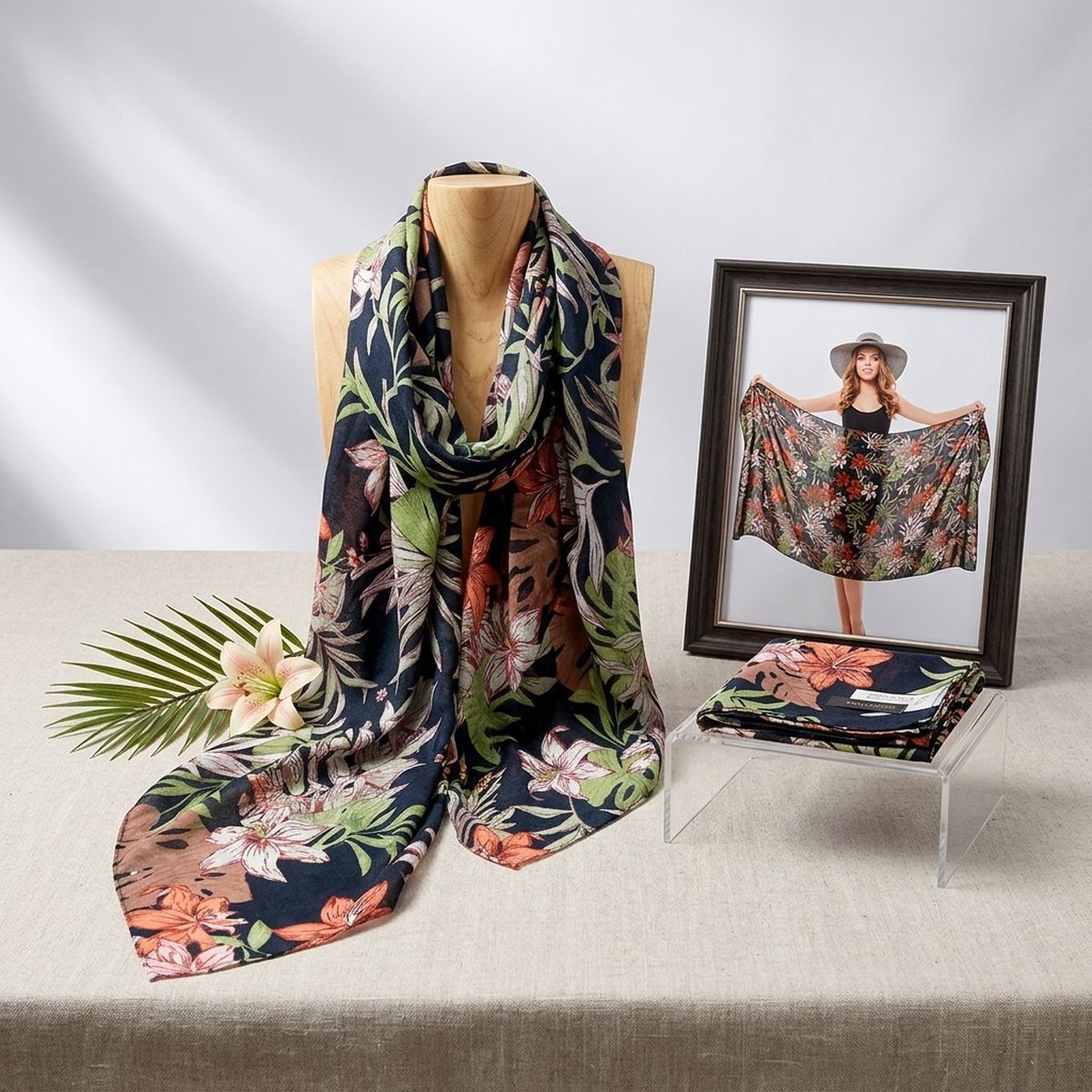 Tropical floral scarf draped around a wooden display bust beside a flower, palm frond, folded scarf, and framed photo of a model holding the wrap open
