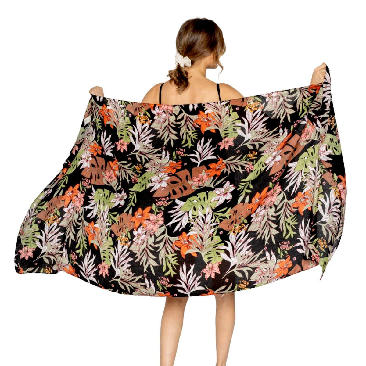 Rear view of a model holding a black tropical floral wrap spread wide, showing green and pale foliage with pink and orange flowers against a white background