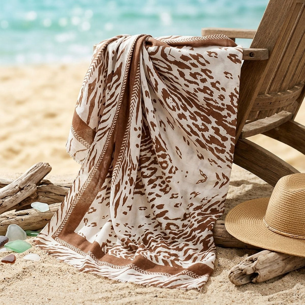 Brown-and-beige animal print wrap draped over a wooden beach chair beside a straw hat, driftwood, and sea glass, with the shoreline blurred behind it.