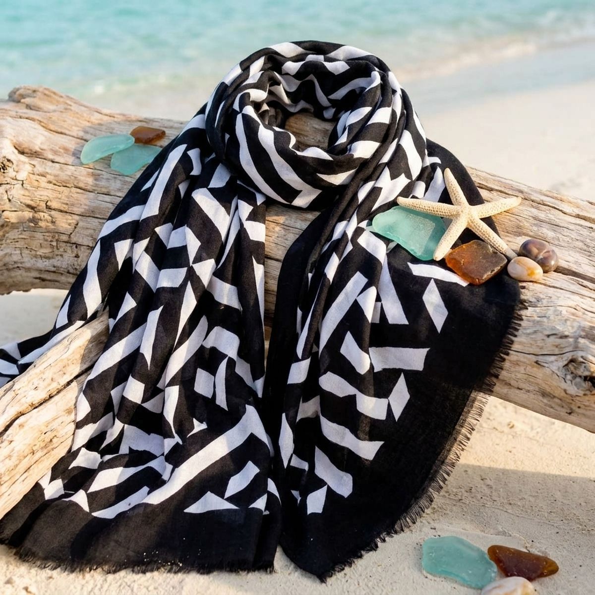 Black-and-white geometric scarf draped over beach driftwood beside a starfish, sea glass, and stones, with turquoise water in the background
