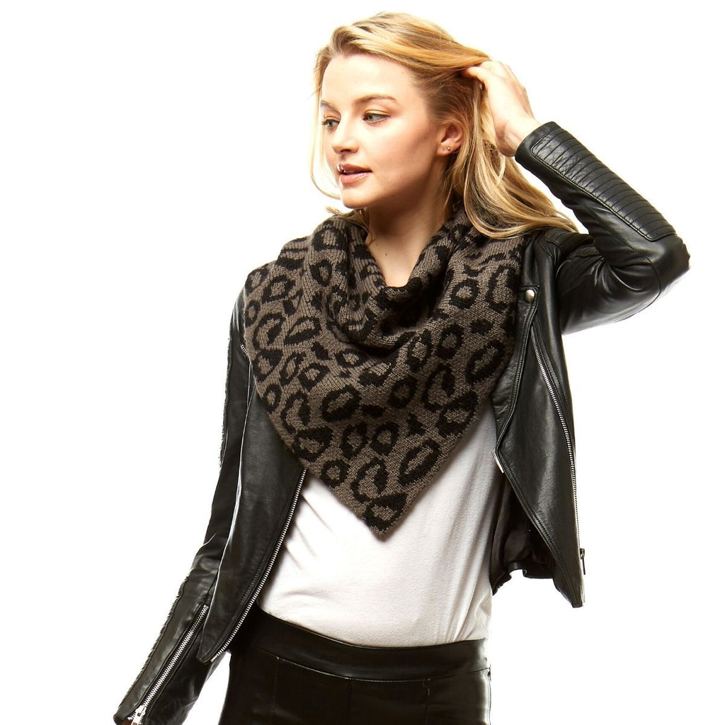 Model lifting her blonde hair while wearing a gray-and-black leopard knit triangle tube scarf over a white top and black zippered biker jacket against a white background