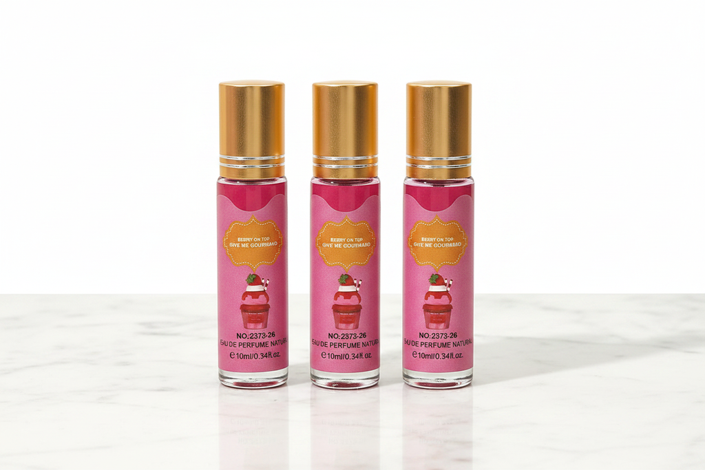 Three 10ml mini eau de parfum roll-on bottles with gold caps, pink and white vertical stripe Berry On Top Give Me Gourmand label with strawberry ice cream illustration, standing upright in a row on white marble surface, white background