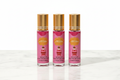 Three 10ml mini eau de parfum roll-on bottles with gold caps, pink and white vertical stripe Berry On Top Give Me Gourmand label with strawberry ice cream illustration, standing upright in a row on white marble surface, white background