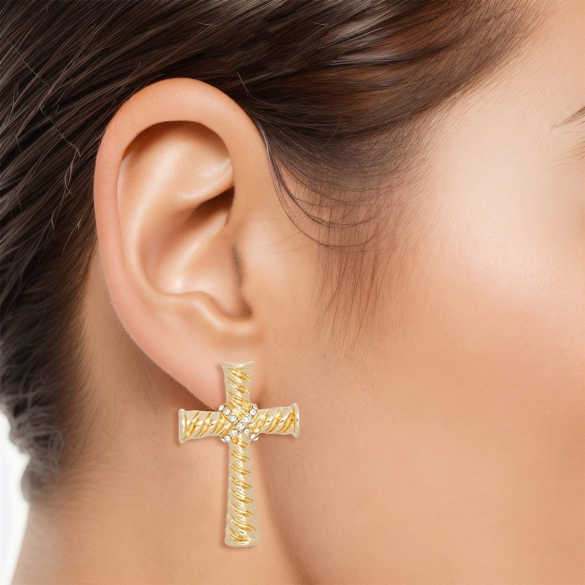 Model wearing a large gold-tone cross stud earring with twisted ridged texture and a clear rhinestone cluster at the center