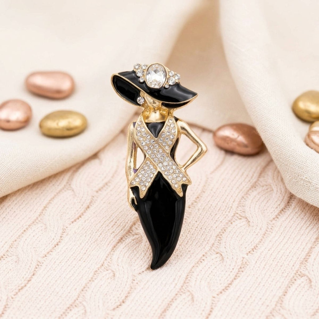 A fashion brooch shaped like a woman in a black and gold hat and dress, embellished with clear rhinestones, displayed on a textured cream knit fabric with decorative rose gold and gold beads.