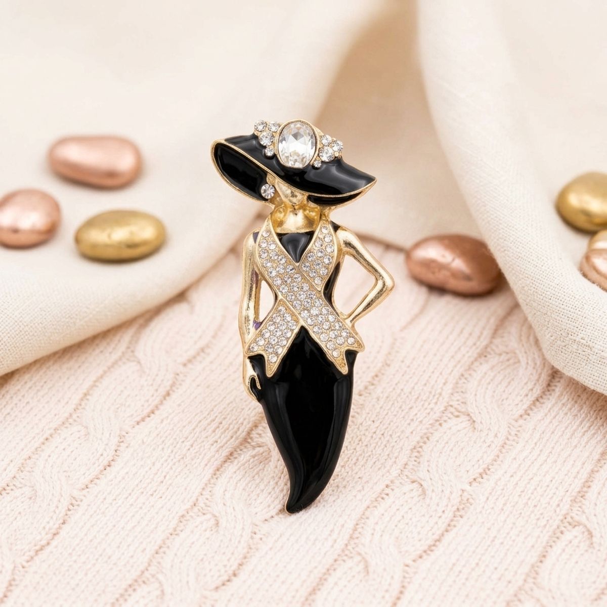 A fashion brooch shaped like a woman in a black and gold hat and dress, embellished with clear rhinestones, displayed on a textured cream knit fabric with decorative rose gold and gold beads.