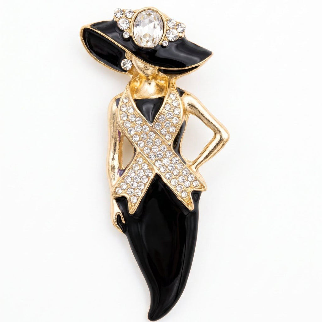 A gold and black enamel fashion brooch shaped like a woman wearing a wide-brimmed hat and a ribbon pin, embellished with clear rhinestones on a white background.