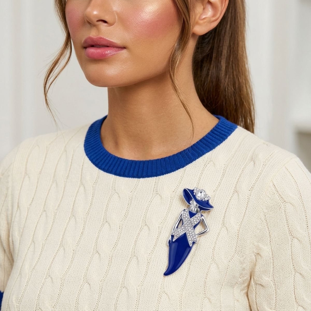A close-up of a woman wearing a cream cable-knit sweater with a royal blue crew neck, accessorized with a blue and silver fashion pin brooch.